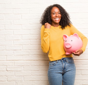 Black women wearing a yellow sweater and jeans with dark brown wavy hair holding a large pink piggy bank
